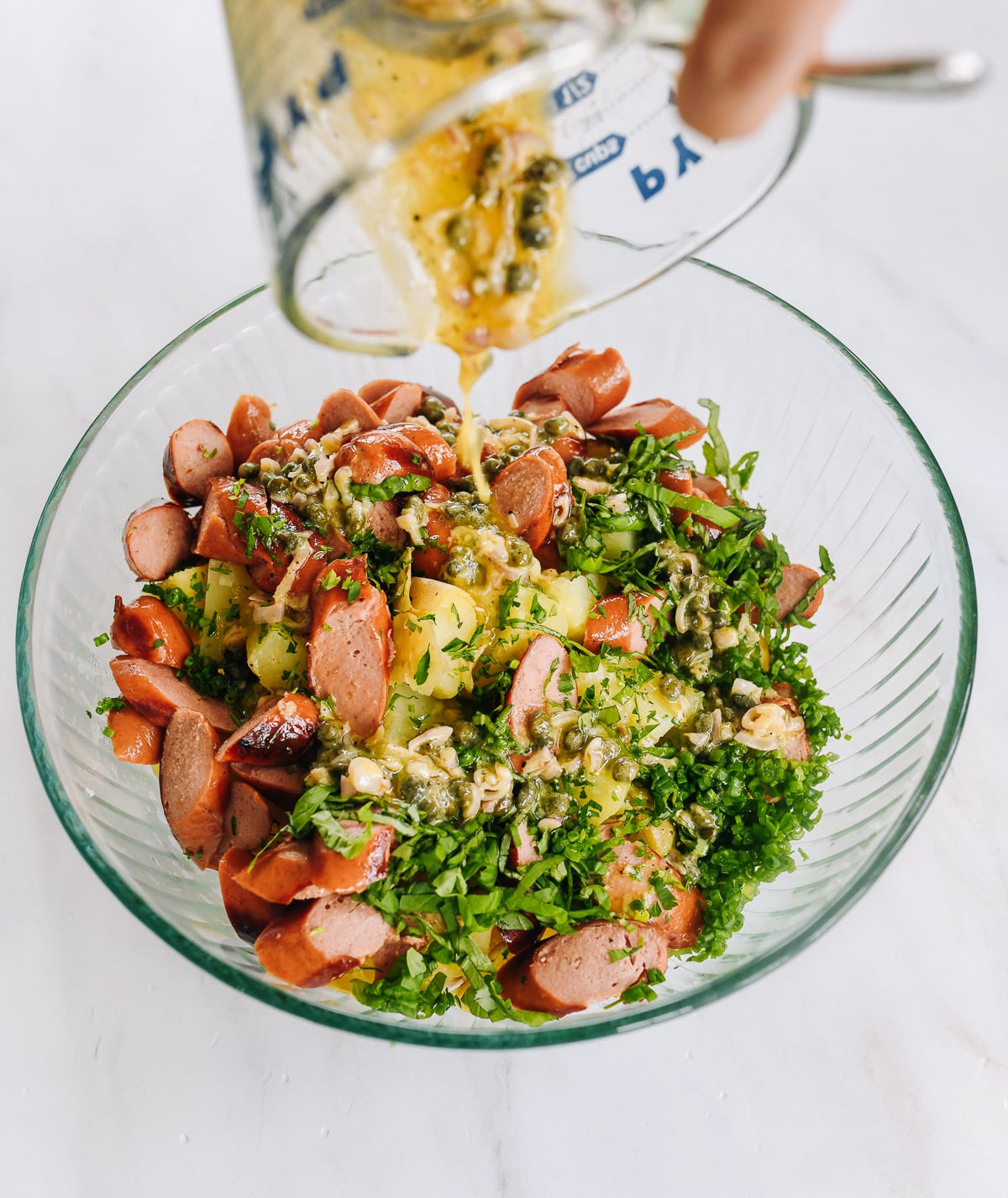 pouring dressing over hot dogs, potatoes, and herbs in glass mixing bowl