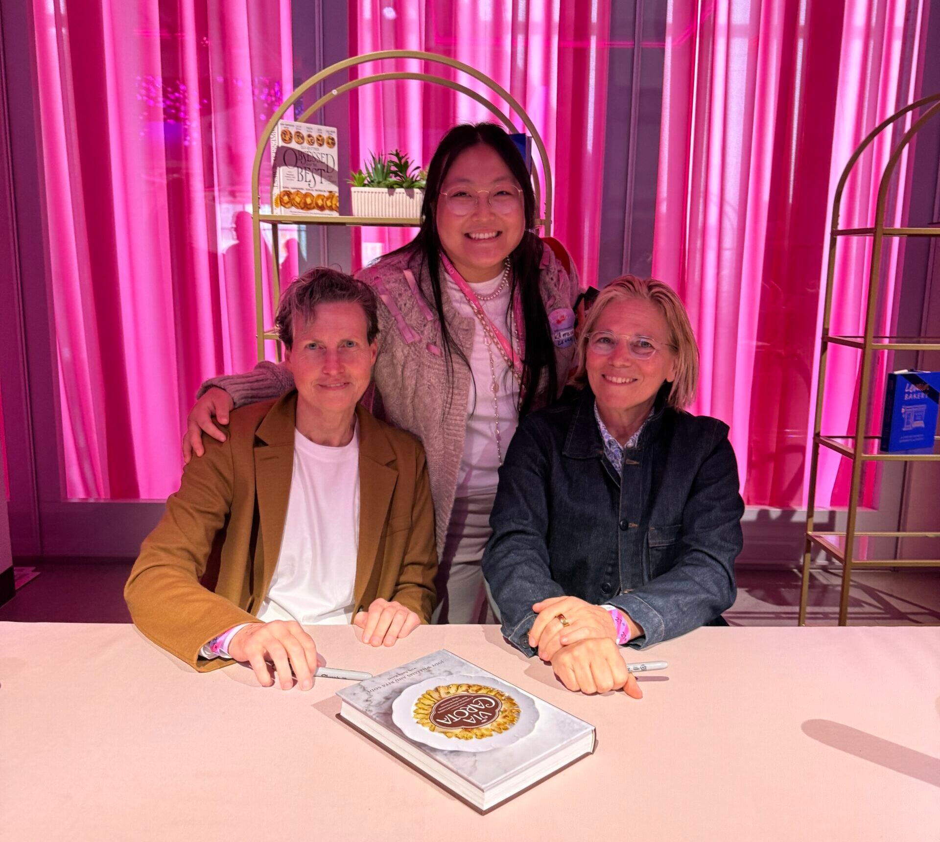 Kaitlin Leung at a book signing with Via Carota chefs Rita Sodi and Jody Williams at a pink table in front of a hot pink wall.
